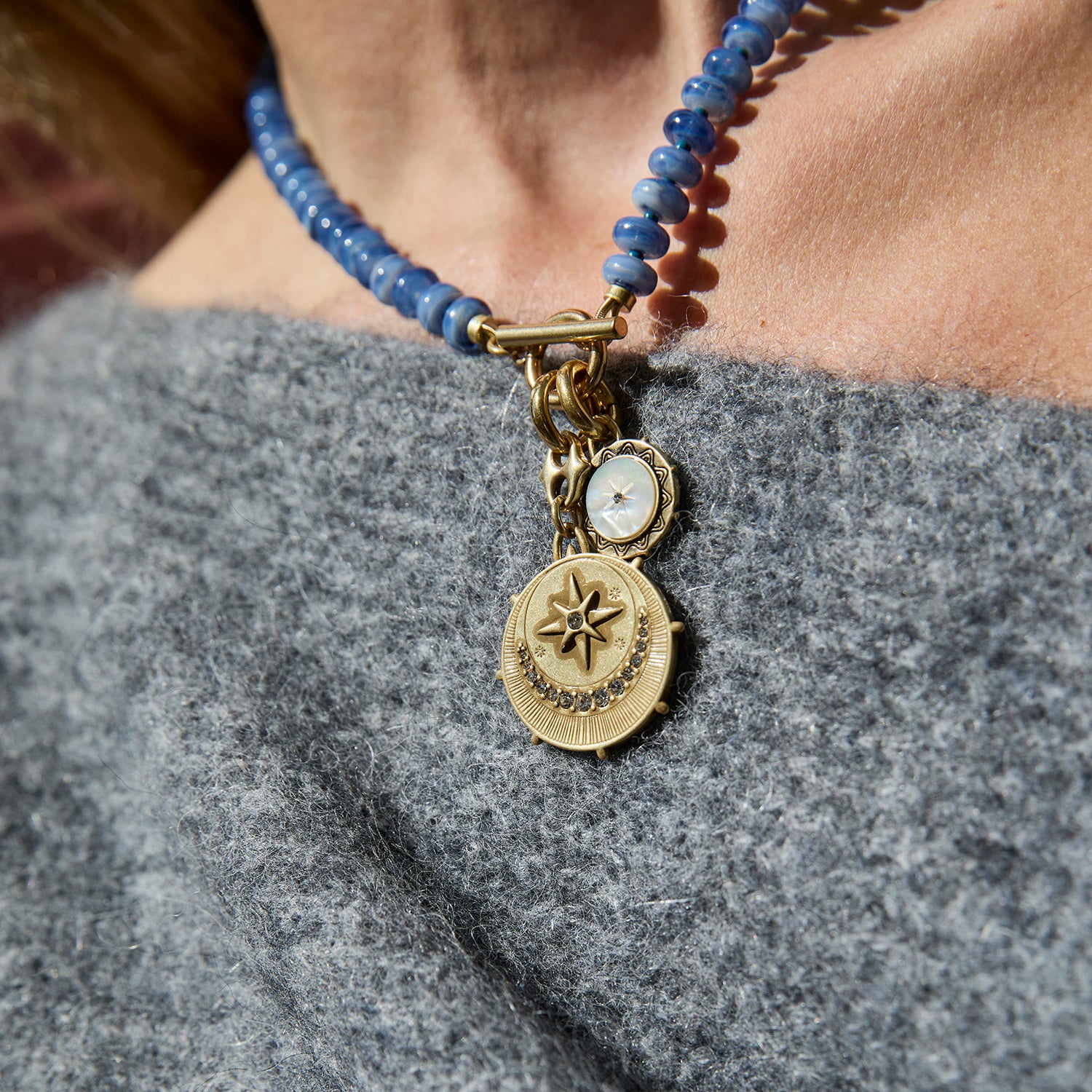 A close-up of a person wearing a beaded necklace with a gold-toned compass pendant and a small round white stone charm, styled over a gray knit top.