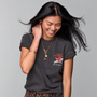 A woman with long black hair smiling and looking down, wearing a dark charcoal gray graphic t-shirt with a red and white 'Tom Petty' graphic on the left chest, a gold pendant necklace, and a gold bracelet, set against a solid gray studio background.