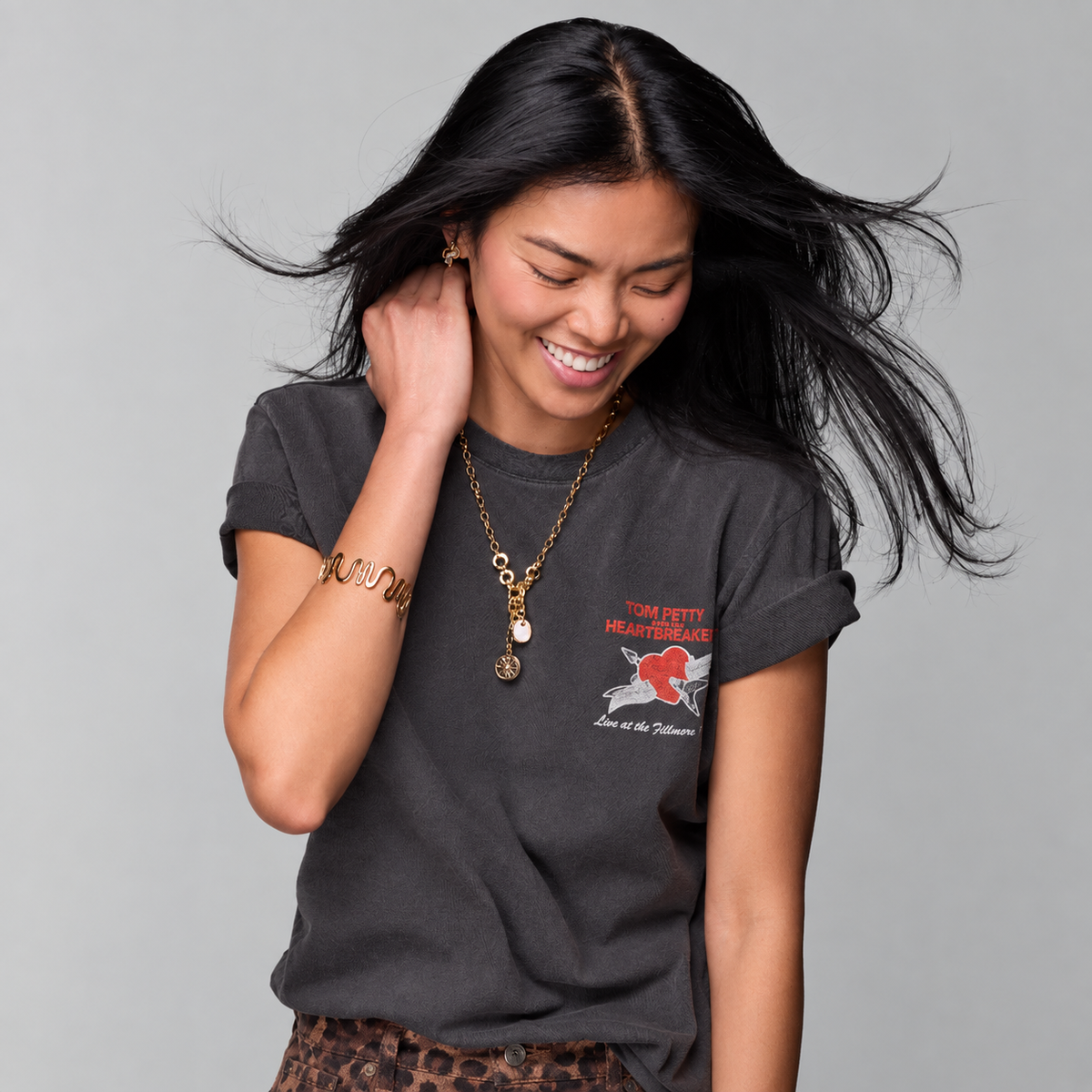 A woman with long black hair smiling and looking down, wearing a dark charcoal gray graphic t-shirt with a red and white 'Tom Petty' graphic on the left chest, a gold pendant necklace, and a gold bracelet, set against a solid gray studio background.