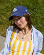 A woman wearing a yellow and white striped tank top, a blue and white striped button-down shirt, a blue New Era baseball cap with an LA logo, and a gold pendant necklace, sitting outdoors on a concrete surface with green grass in the background.