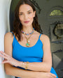 A woman wearing a bright blue tank top and layered gold jewelry, including a green pendant necklace and a beaded choker, posing in front of a dark decorative door.