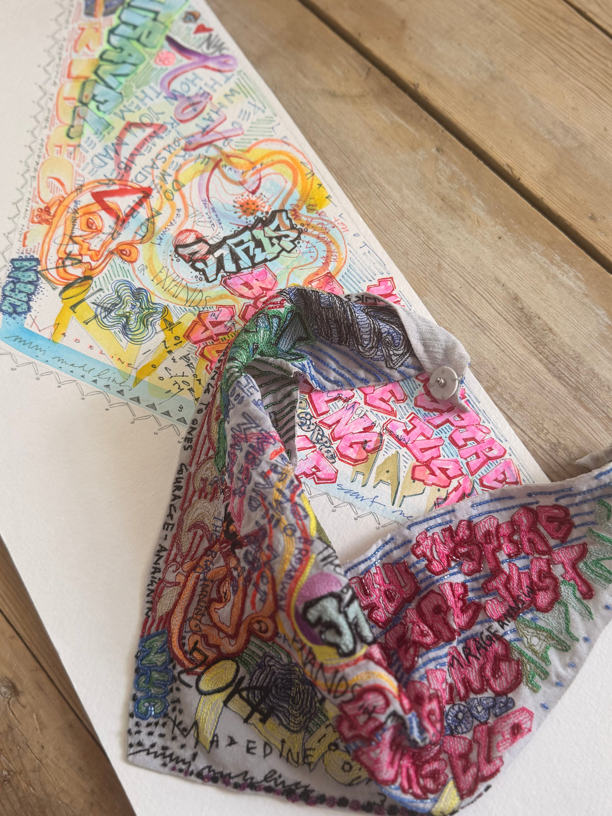 Sketch with colored graffiti next to an embroidered scarf necklace on a wooden table.