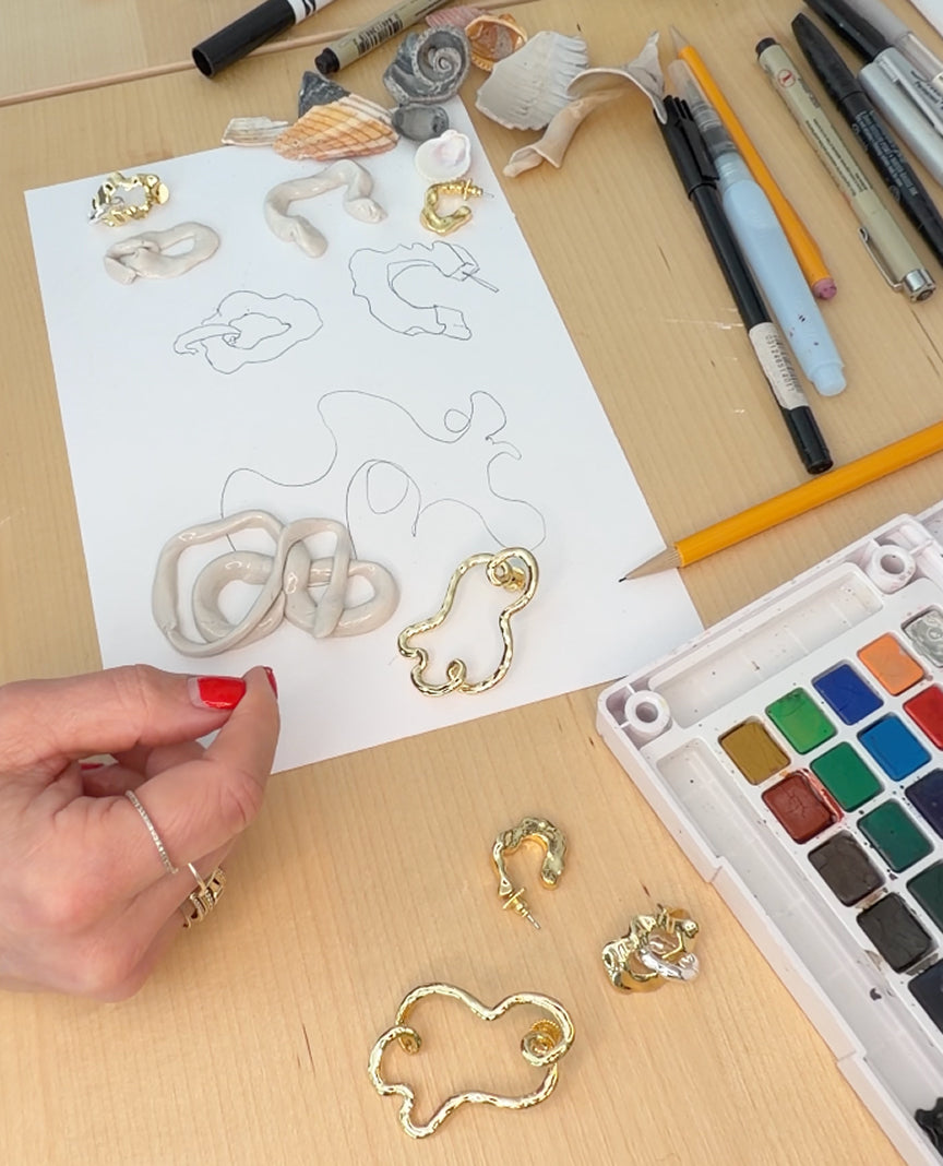 Hand holding gold jewelry piece with design sketches on paper and art supplies on a table