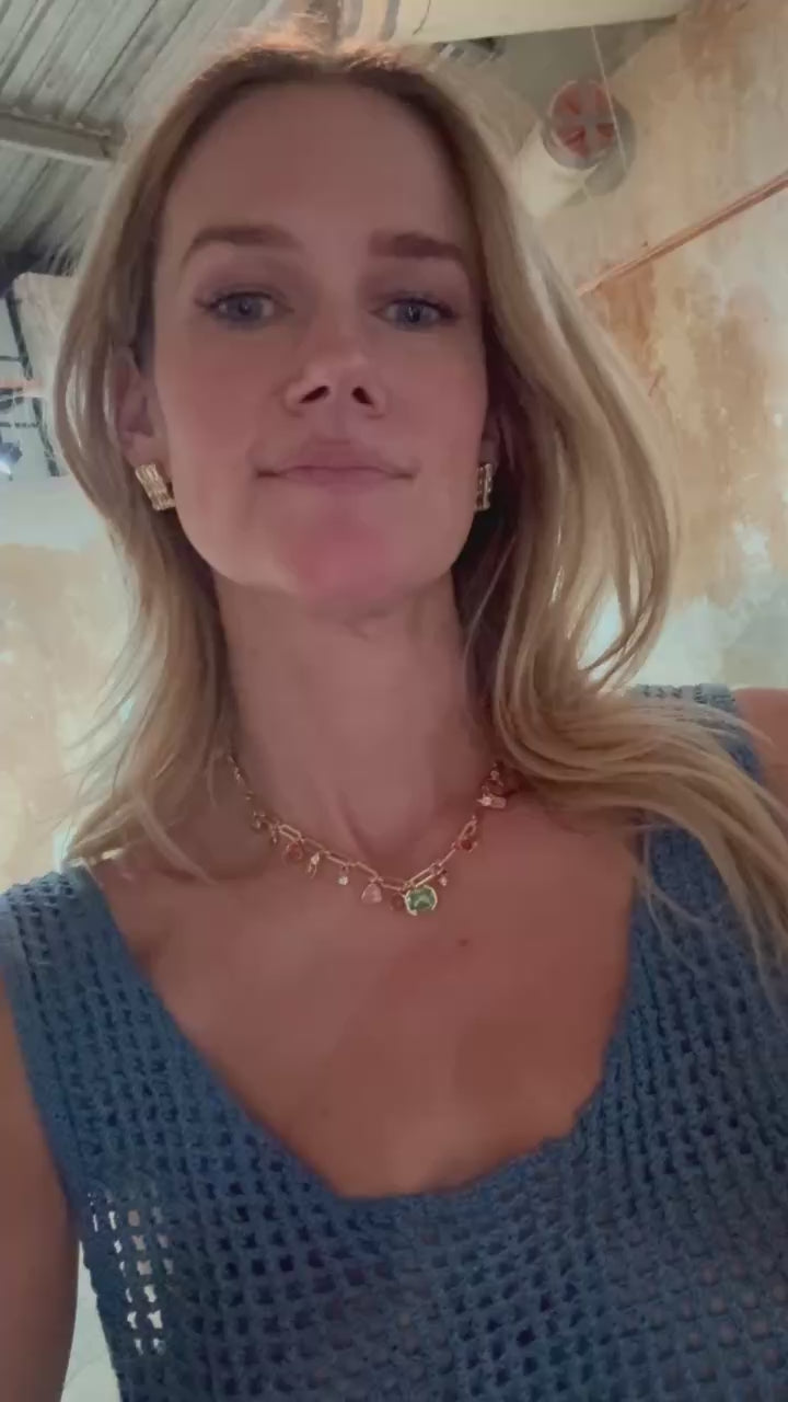 Model wearing colorful gemstone charm necklace and gold and crystal stud earrings wearing a blue dress against a stone background