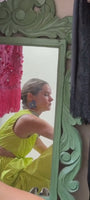 Model wearing blue floral shaped earring made with beads and threads in a yellow dress looking into a mirror 