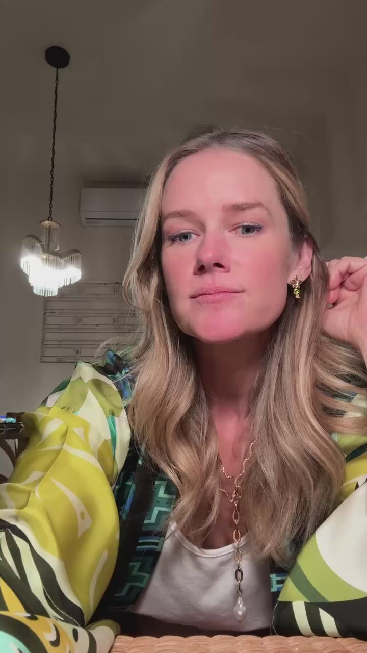 Model showing off gold palm tree shaped stud earring with a long gold chain necklace with a pearl pendant wearing a multicolor robe