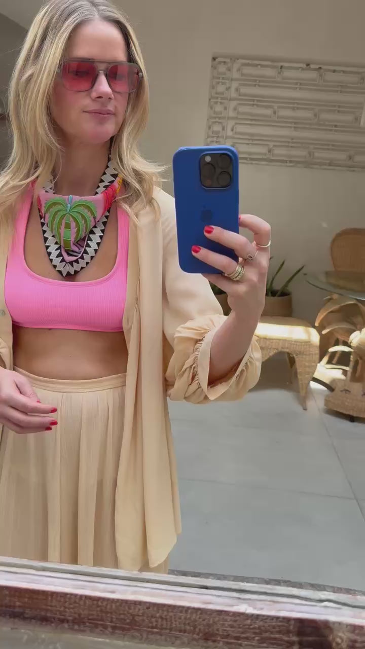 Model wearing peach cover up set with pink swimsuit tying a colorful embroidered and beaded scarf necklace around neck with sunglasses on