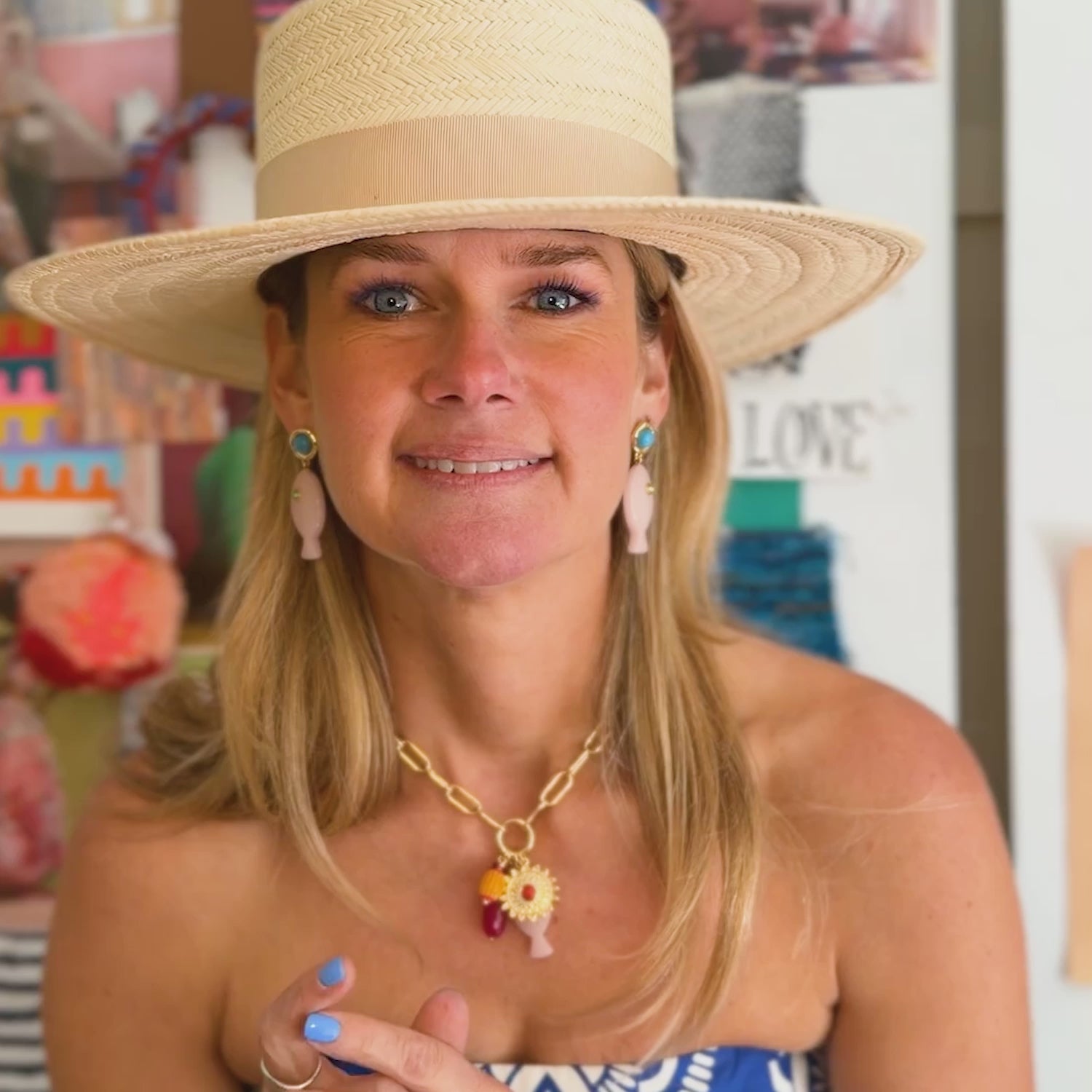 Model wearing a straw hat and blue dress while explaining the design begin the Liliana Charm earrings, pink fish shaped earrings with a blue stone topper