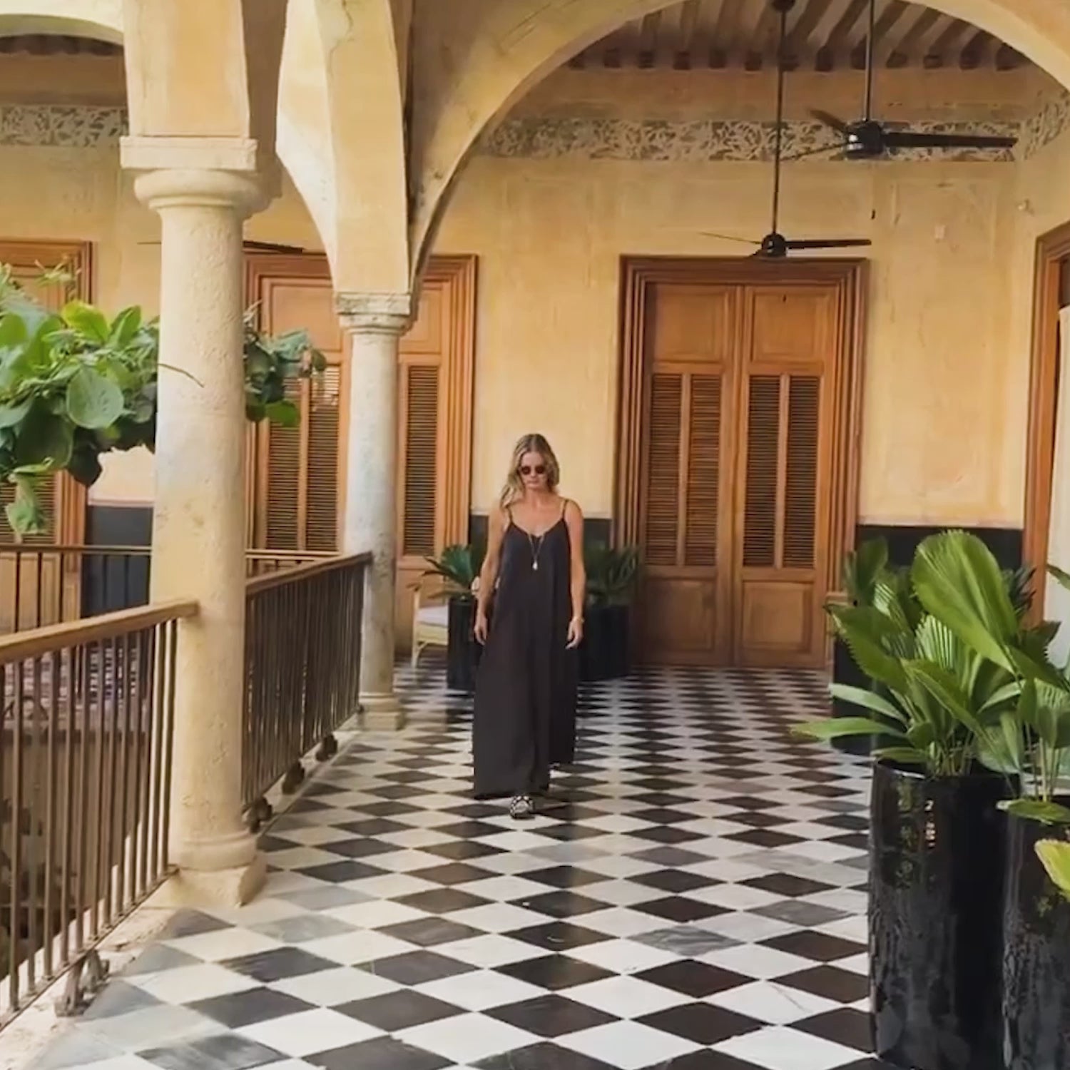Woman walking down a hallway wearing a black dress with long chain gold necklace with a shell pendant