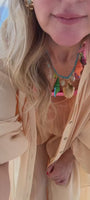Model wearing peach colored top with pink bandeau underneath. Showing off a shell and semi precious stone necklace around her neck with gold drop earrings on the streets of Merida, Mexico