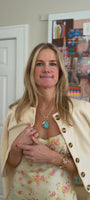 Model explaining a turquoise shell shaped drop necklace with gold chain while wearing a yellow dress and ivory sweater on shoulders