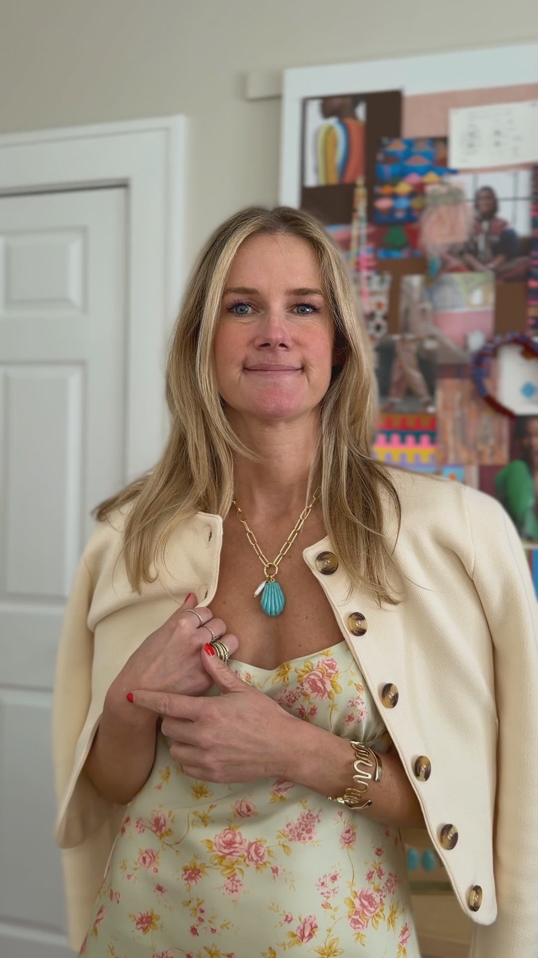 Model explaining a turquoise shell shaped drop necklace with gold chain while wearing a yellow dress and ivory sweater on shoulders