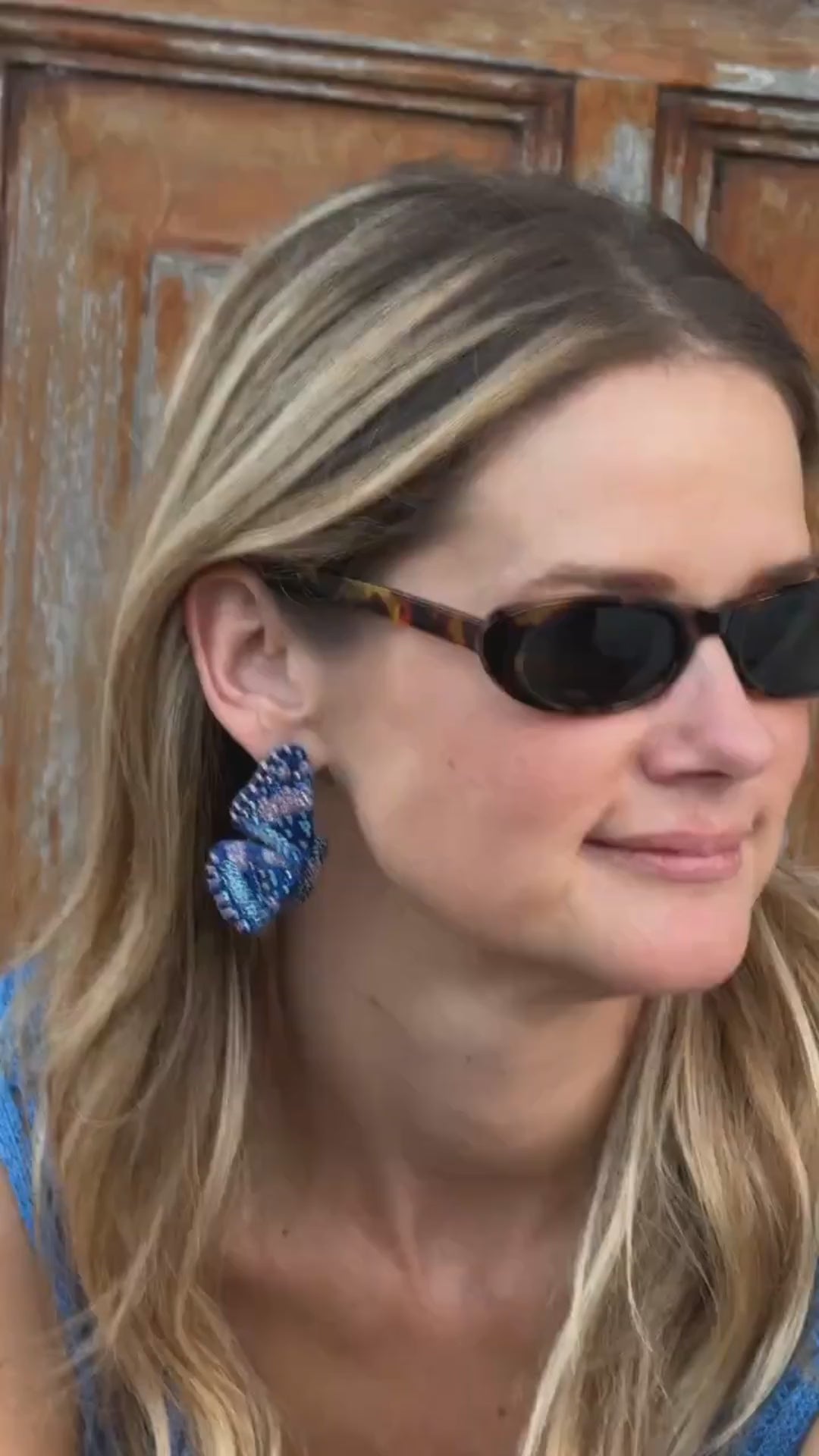 Model wearing beaded and embroidered butterfly earrings with sunglasses on her face against a wooden door