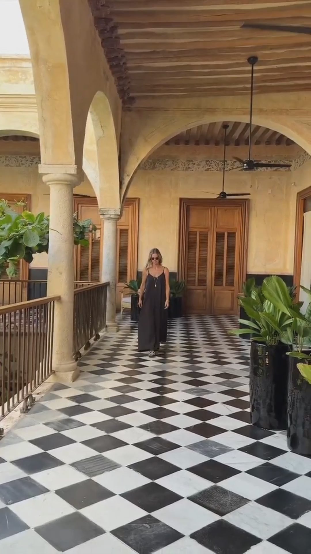Model wearing long black dress walking down a hallway with a long gold chain necklace and shell pendant