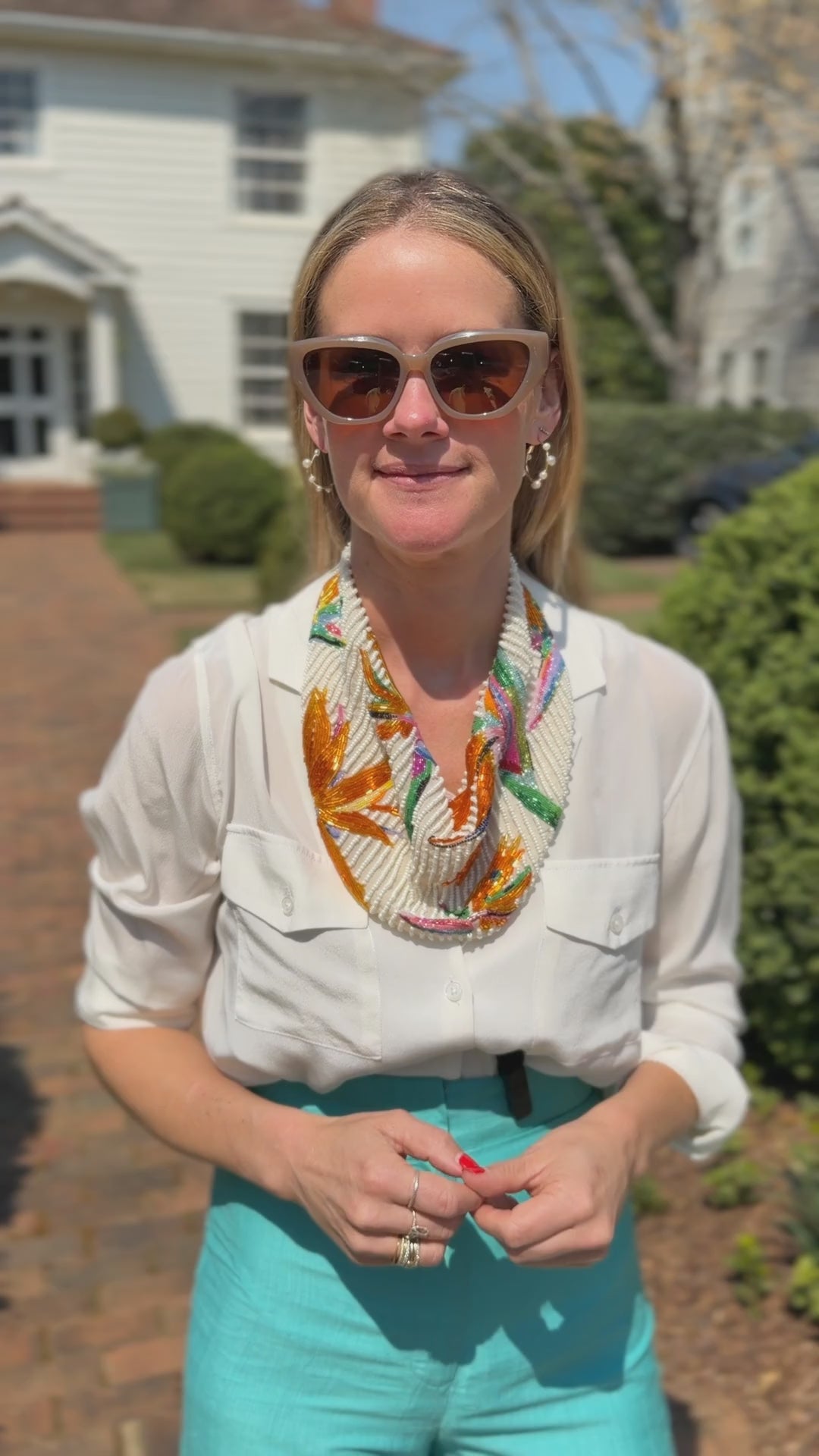 Video of model wearing colorful beaded scarf necklace outside a white house in a white top and blue pants