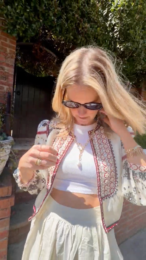 Model wearing bolero with white skirt explaining gold chain necklace with pearl drop charm outside