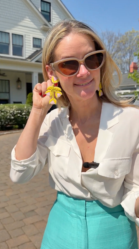 Model standing in front of a house wearing yellow tulip shaped earrings in a white blouse and teal bottoms