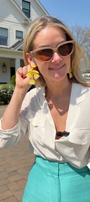 Model standing in front of a house wearing yellow tulip shaped earrings in a white blouse and teal bottoms