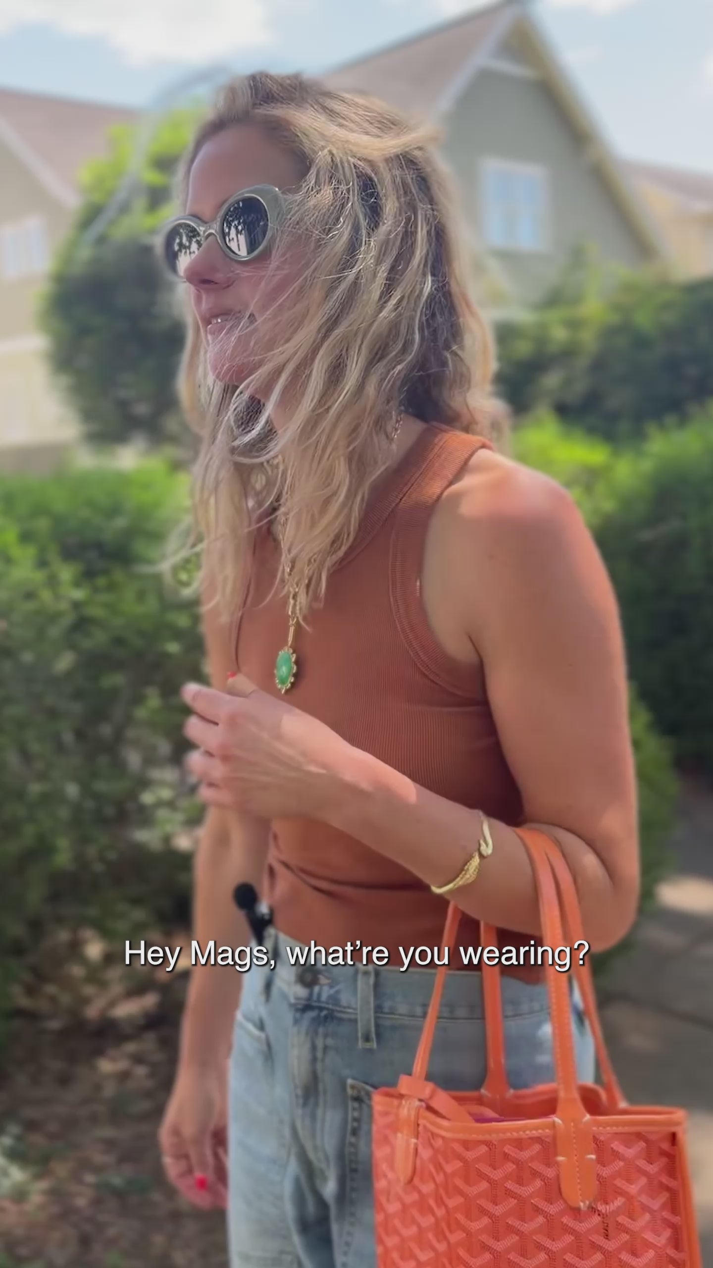 Model walking and wearing a gold chain necklace with a green pendant attached with a tote in her hand and sunglasses on