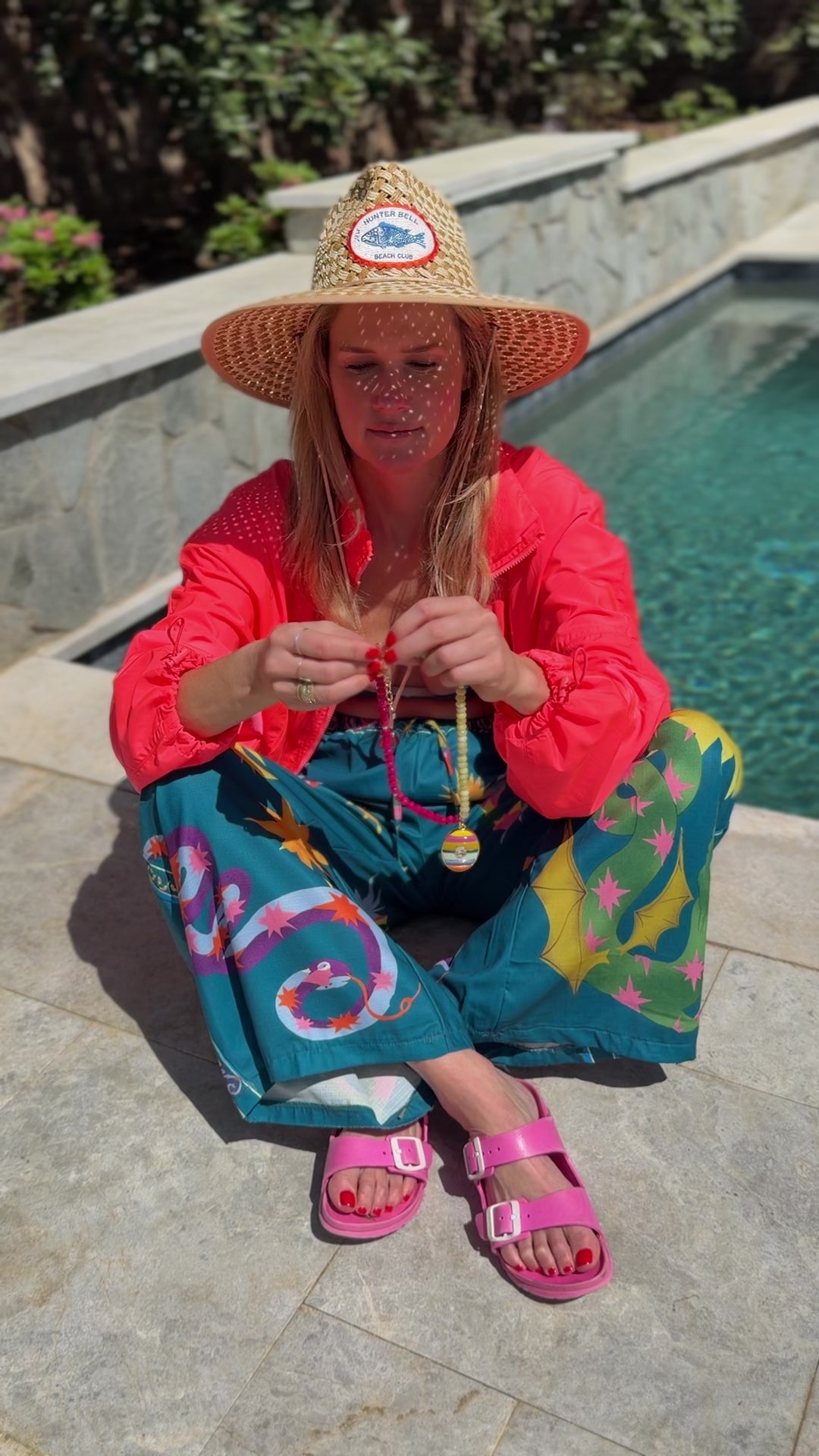 Model sitting next to a pool wearing a swim coverup and straw hat showing a pink and yellow beaded necklace with a colorful pendant