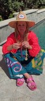 Model sitting next to a pool wearing a swim coverup and straw hat showing a pink and yellow beaded necklace with a colorful pendant