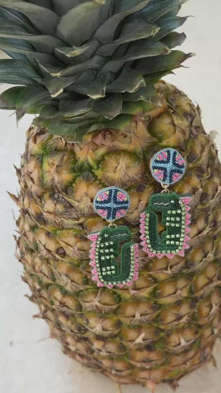 Alligator shaped earrings with green blue and pink accents resting on a pineapple against a white background