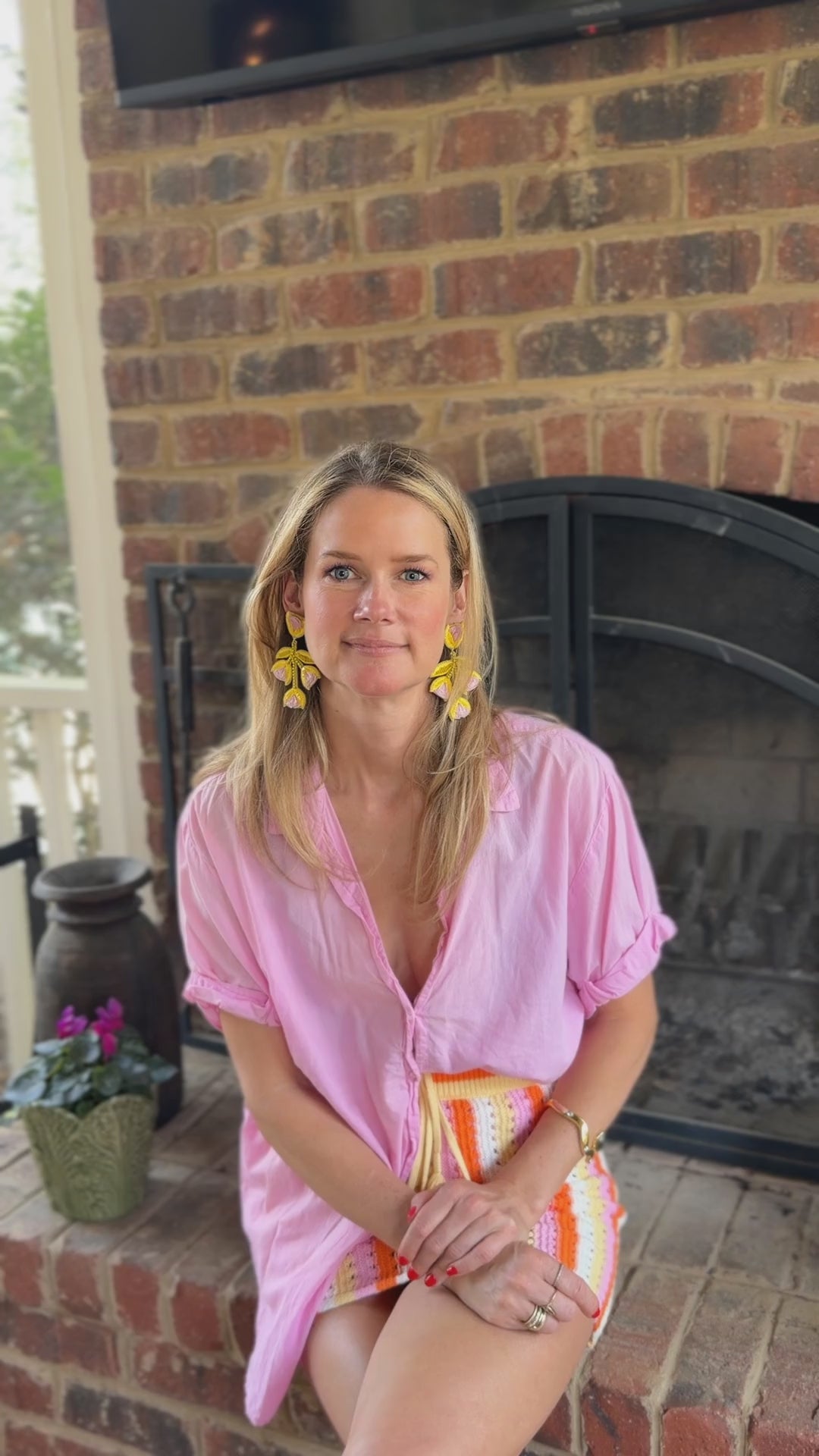 Model wearing yellow tulip shaped earrings in pink blouse and knit skirt against a fireplace